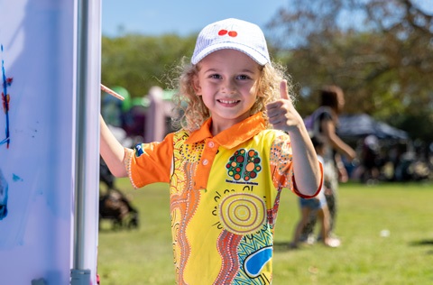A young child giving a thumbs up at an outdoor event.